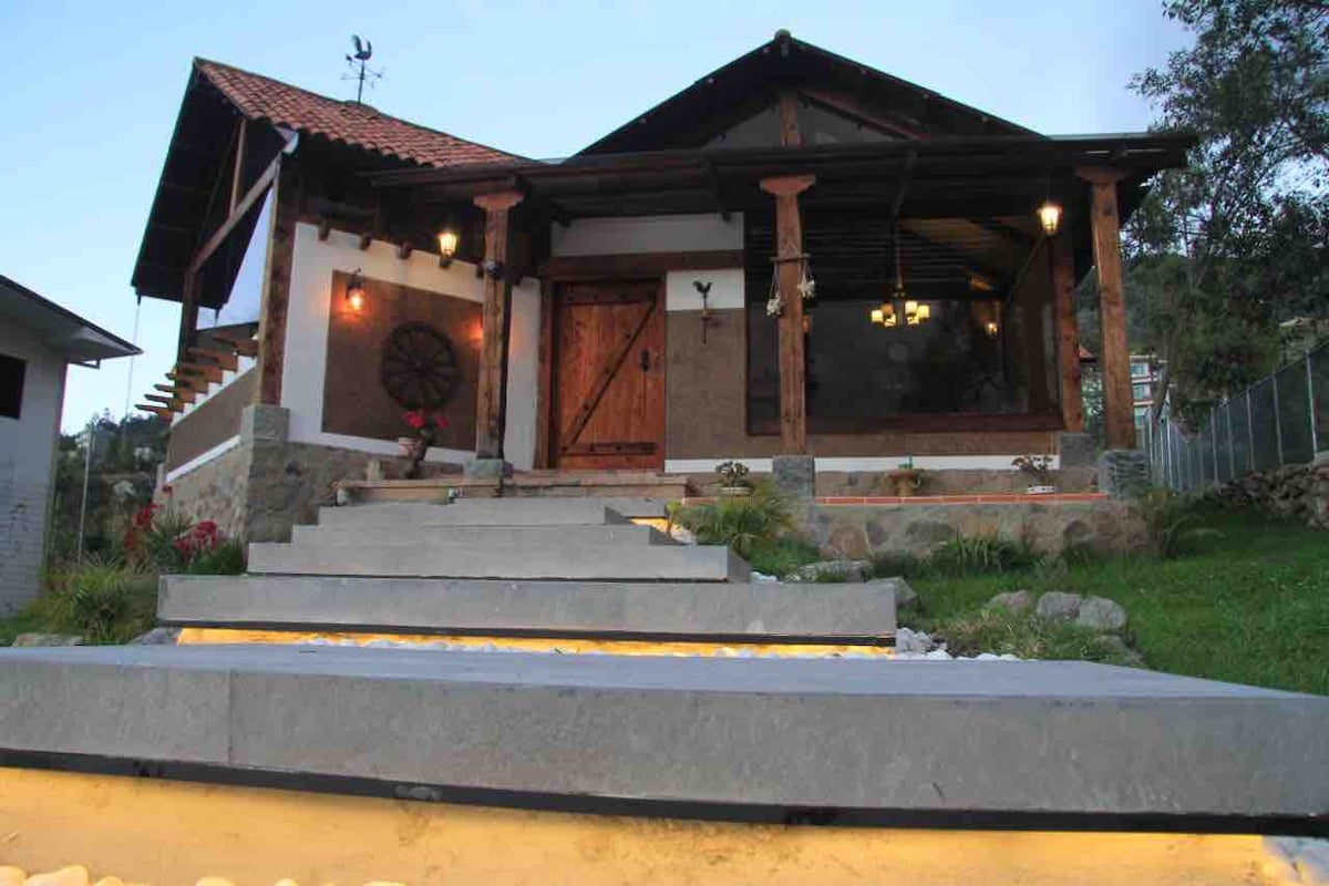 The villa features a well-defined entrance with stone steps leading up to a wooden door framed by large windows. The exterior showcases a combination of stone and wood, punctuated by decorative lighting and landscaped areas adorned with plants and stones.