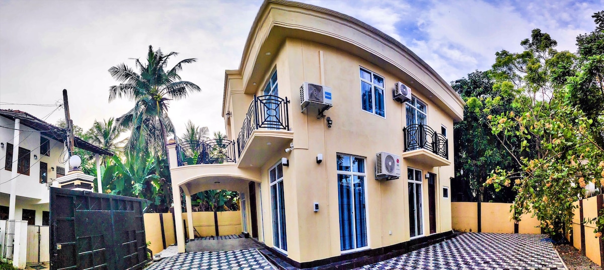 The two-story house features large glass windows and a spacious exterior with a patterned tile surface. Lush greenery and palm trees surround the property, creating a sense of openness and connection to nature.