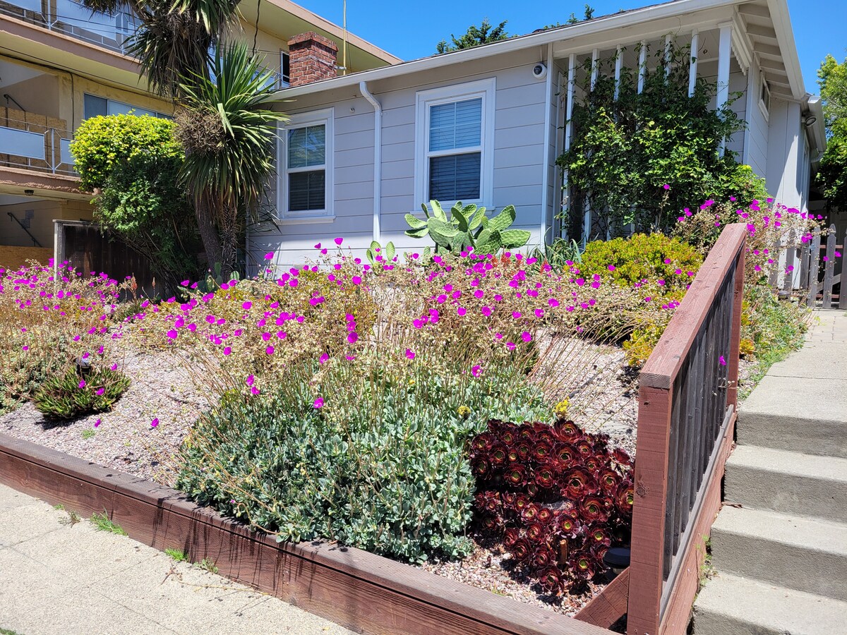 The exterior of the duplex features a well-maintained front garden filled with vibrant purple flowers and green succulent plants. A wooden raised garden bed enhances the landscaping, while a clear blue sky serves as a backdrop to this inviting entryway.