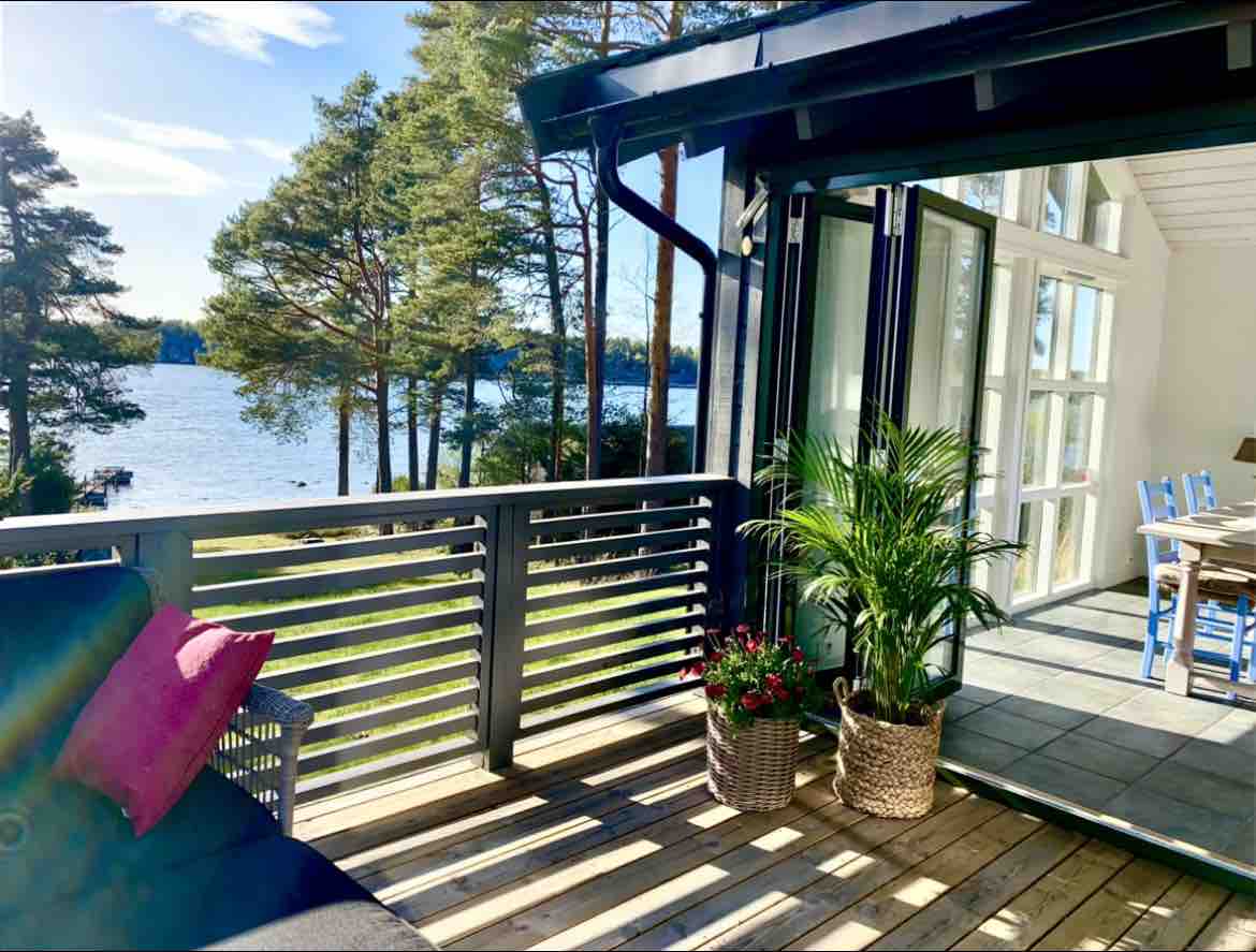 A spacious balcony offers a view of the serene water, framed by trees. Indoor and outdoor living is enhanced by large glass doors, allowing natural light to fill the area. Potted plants and floral arrangements add a touch of greenery to the inviting space.