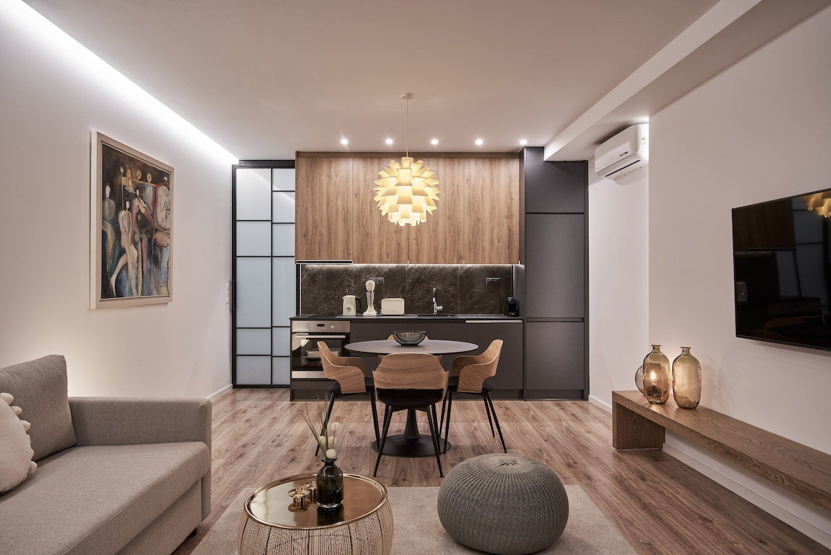 A modern living area features a cozy sofa and a round coffee table. A stylish dining table with four chairs is positioned adjacent to a sleek kitchen, equipped with contemporary appliances. Warm ambient lighting is provided by a unique pendant light, enhancing the elegant decor.