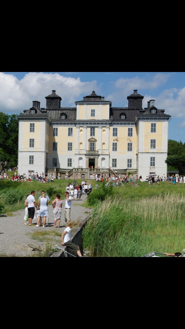 A grand historic building is depicted, showcasing ornate architecture with tall windows and a symmetrical design. Surrounding the structure, a group of visitors is seen, along with lush greenery and a pathway leading to the entrance. The sky above is bright with scattered clouds.
