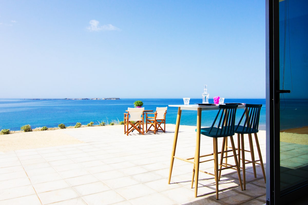 A spacious outdoor terrace features a high table with two stools, offering an unobstructed view of the sea. Additional seating is provided by a set of four wooden chairs nearby. The clear blue sky complements the serene ocean landscape, creating a calming atmosphere.