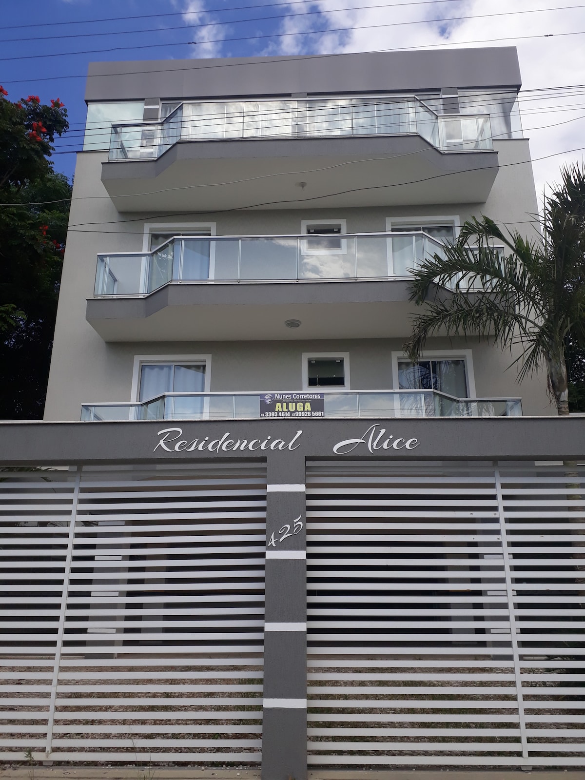 A modern three-story building features a light-colored façade with large balconies on each level. The entrance is marked by a stylish sign reading 'Residencial Alice.' A decorative fence provides privacy, while tropical plants add a touch of greenery to the exterior.