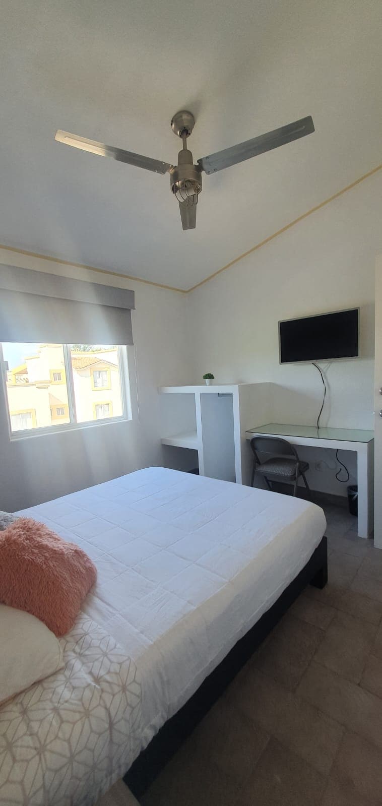 A bright bedroom features a spacious bed with a soft white blanket. Natural light enters through a large window, illuminating the room. A desk and chair are positioned against the wall, and a small television is mounted nearby. A ceiling fan adds to the comfort.