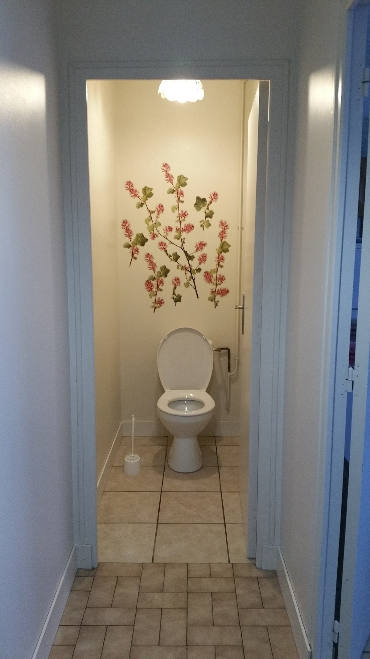 A well-lit bathroom is depicted with light-colored walls and tiled flooring. Floral wall art adds a decorative touch. The image shows a standard toilet positioned at the back, with a toilet brush visible on the floor nearby. Interior space appears neat and organized.