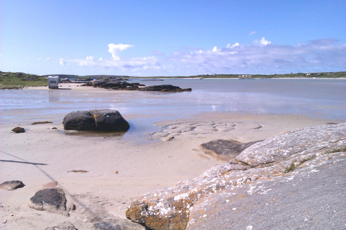 A serene beach landscape features smooth sand and exposed rocks at low tide. Gentle waves can be seen lapping at the shore, with a clear blue sky overhead dotted with clouds. The coastline stretches into the distance, offering a peaceful, natural setting.