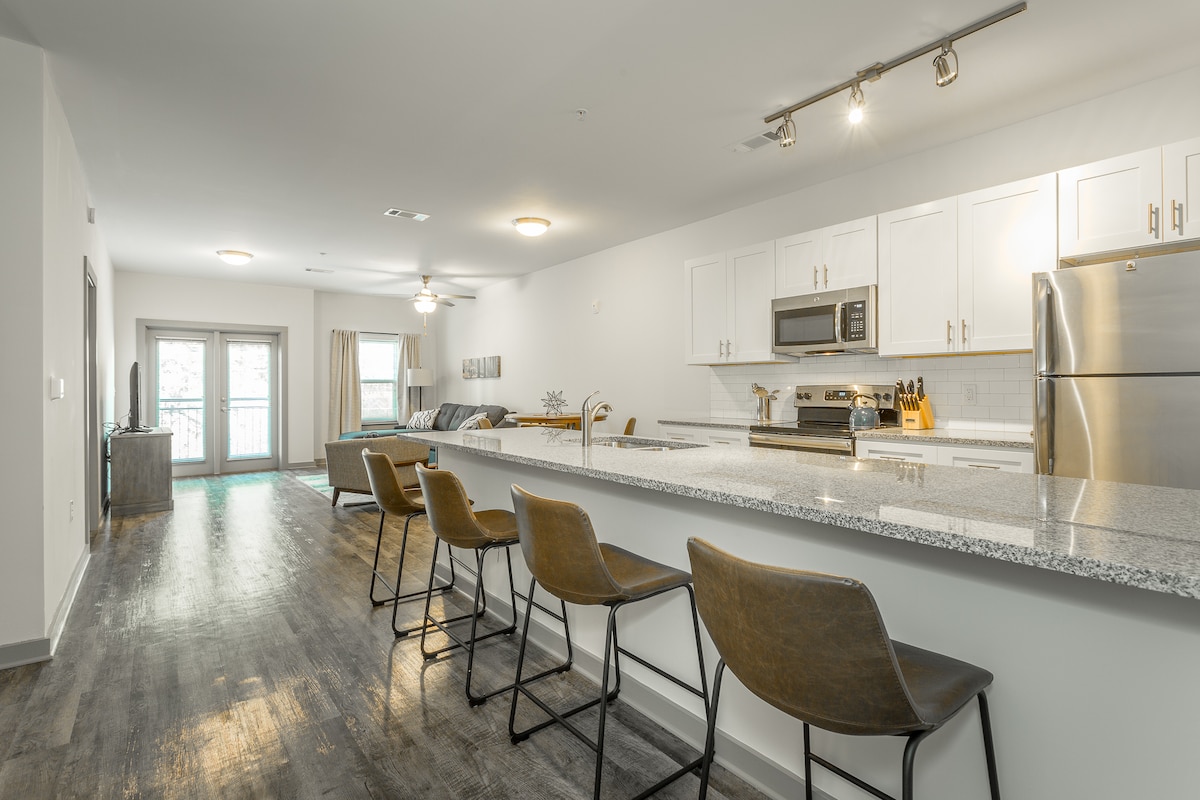 A modern kitchen area features sleek white cabinetry and stainless steel appliances, complemented by a granite countertop. Four barstools line the counter, inviting casual seating. The open layout leads to a living space with natural light flowing through sliding glass doors, revealing a view of a balcony.