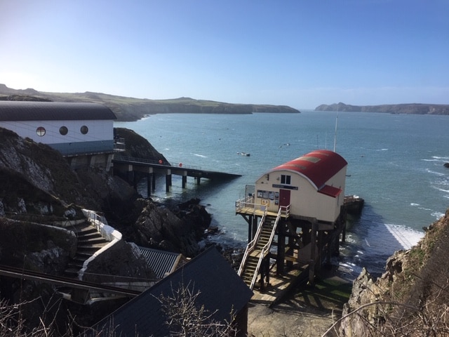 The image captures a scenic coastal view featuring two structures on stilts adjacent to the water. The shoreline is rocky, with gentle waves lapping at the base. The sky is clear, showcasing bright blue hues, while distant hills create a tranquil backdrop.