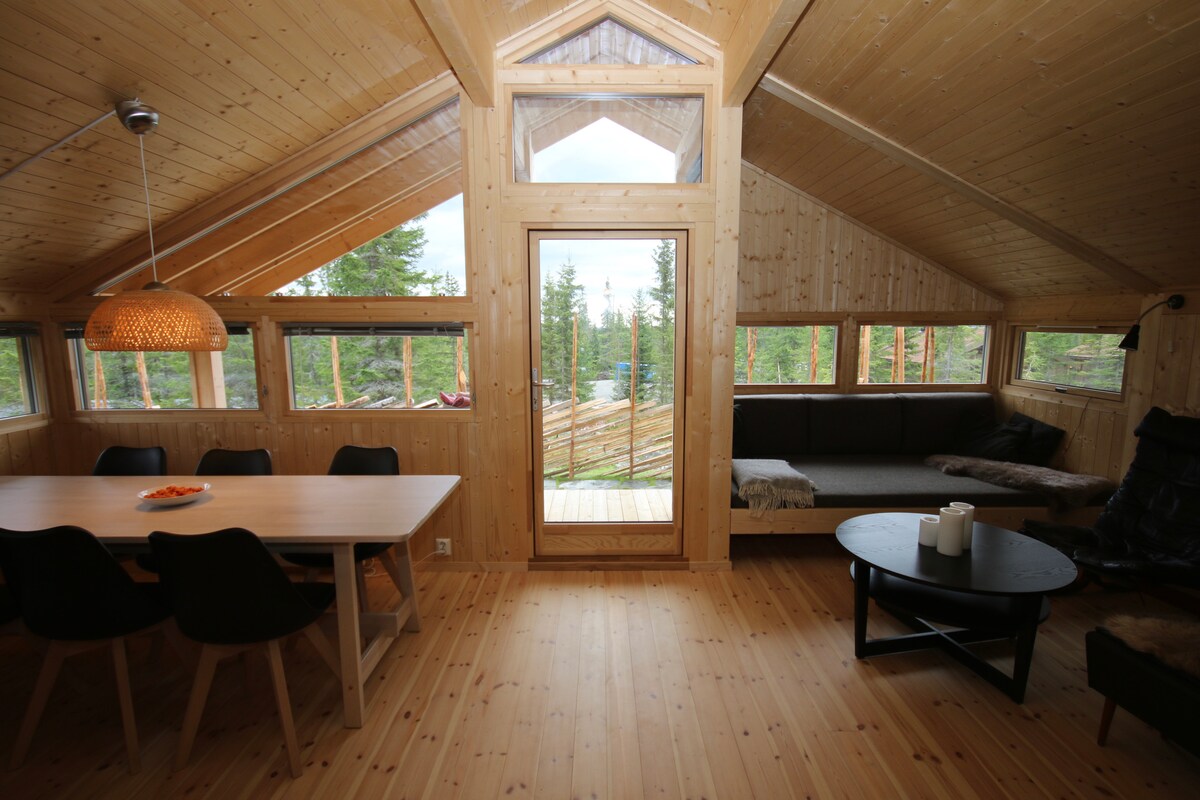 An open living area with wooden walls and ceilings is illuminated by natural light through large windows. A light wood dining table with black chairs is visible near a cozy seating area featuring a dark couch and a round coffee table. A glass door leads to an outdoor deck.