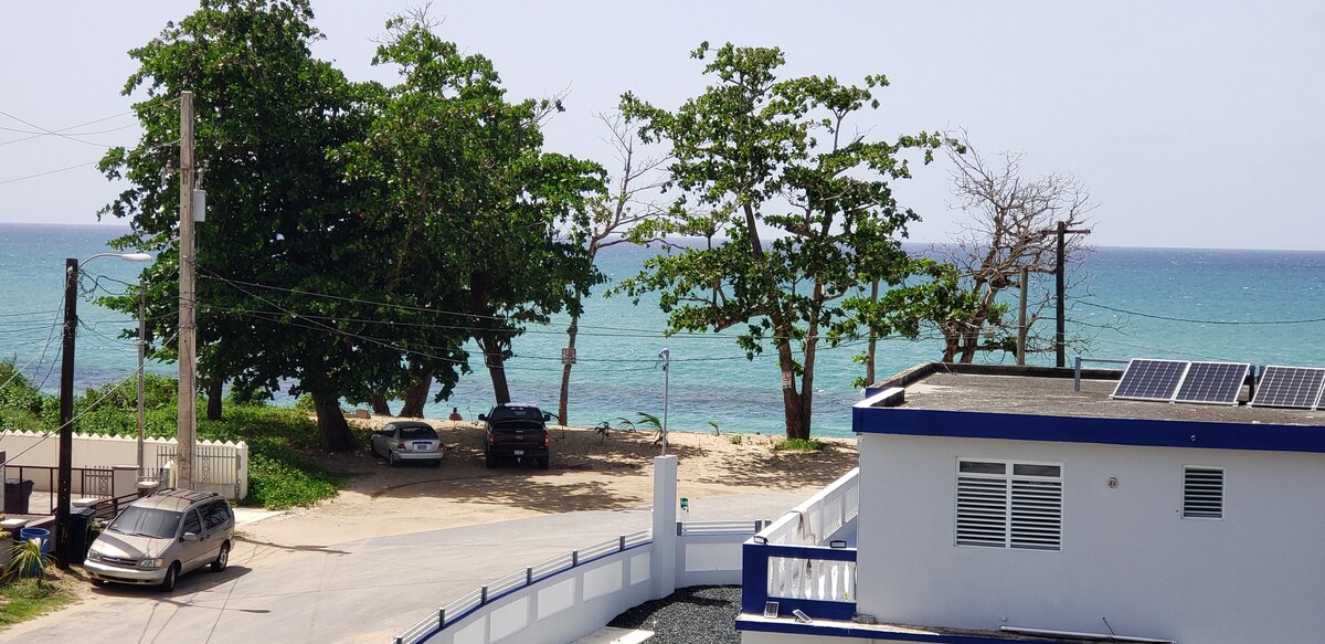 A scenic view showcases the ocean with varying shades of blue, framed by lush green trees. The coastline is visible, with vehicles parked nearby, and the charming apartment building occupies the foreground, accented by solar panels on the roof.