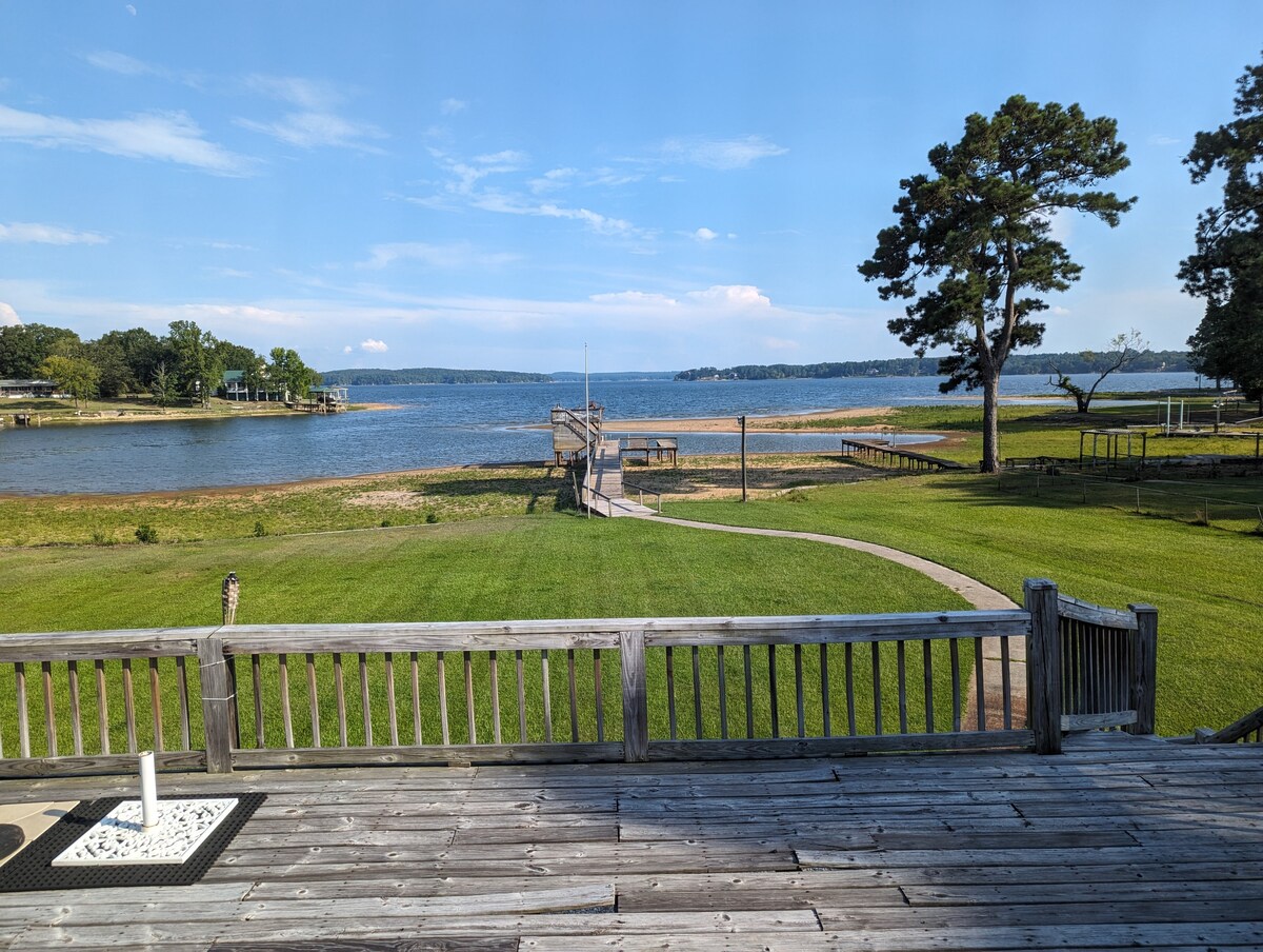 A wide view showcases the lake and surrounding landscape from the deck. The grassy area leads towards a private dock, extending into the water with a path running alongside. The scene is completed by trees lining the shore and a clear blue sky overhead.