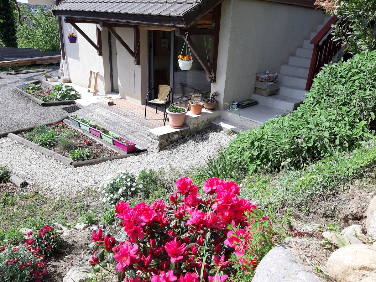 The entrance to a freshly renovated apartment is framed by vibrant flowers and plants. A well-maintained path leads to the door, with potted plants adorning the entryway. The surrounding area includes a mix of gravel and greenery, creating an inviting outdoor space.