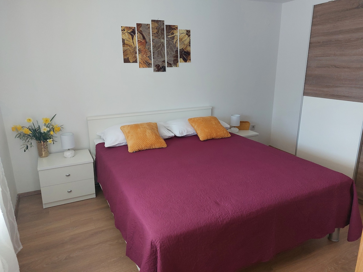 A sleeping area features a spacious bed covered with a deep red bedspread. Two orange cushions are positioned at the head of the bed, complemented by bedside tables with lamps. A wall display of abstract art is mounted above the bed, while a vase of yellow flowers adds a touch of color.