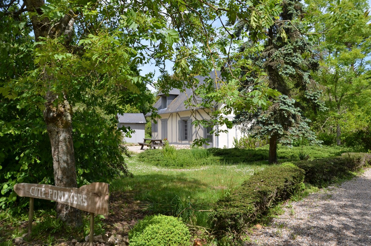 A charming independent house is nestled within a green landscape, surrounded by trees and grassy areas. A sign reading 'Côté Pommiers' is visible in the foreground, indicating the entrance. The exterior features a sloped roof and multiple windows that create a welcoming appearance.