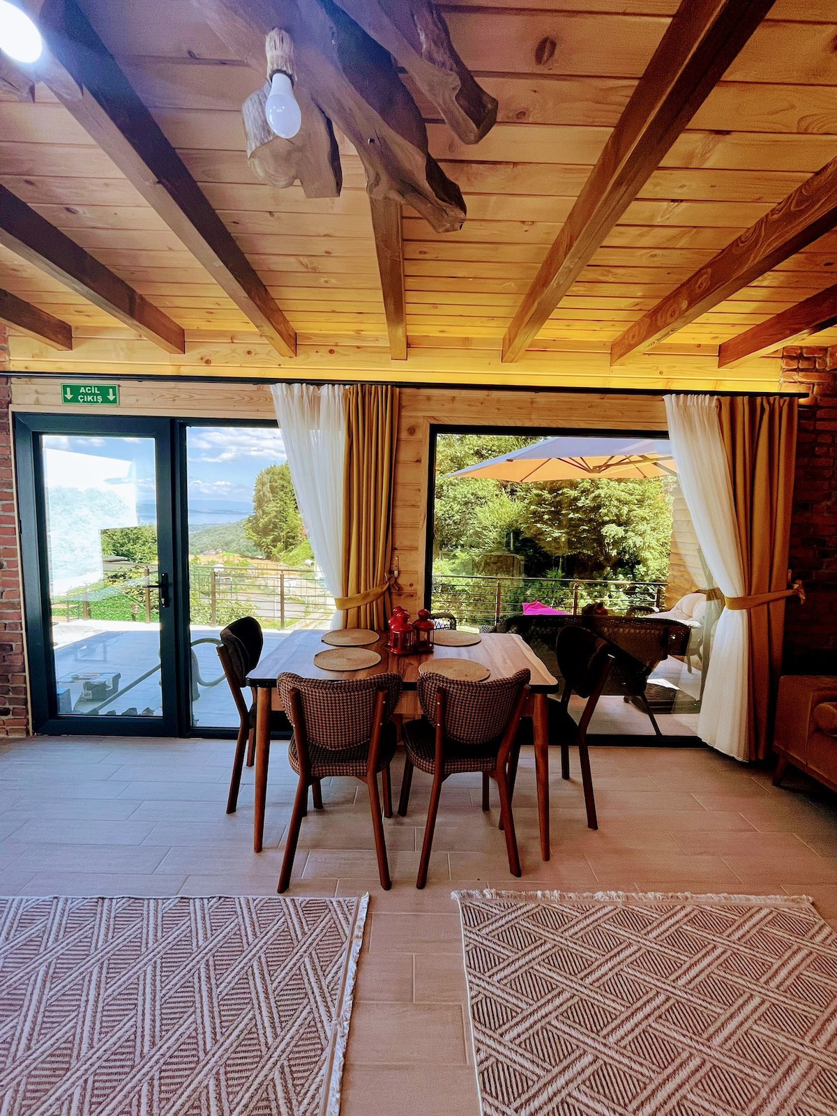 A dining area is photographed, featuring a round table surrounded by four wicker chairs. Large glass doors offer views of the outdoor space, complemented by curtains and light wooden beams overhead. Neutral-toned rugs enhance the inviting atmosphere.