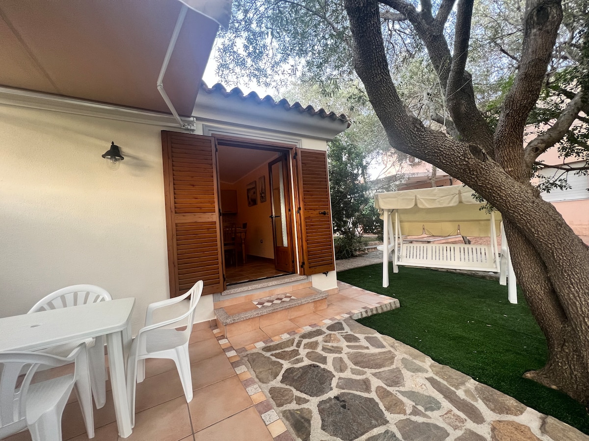An outdoor area is seen with a light-colored building featuring wooden shutters. A small table with two chairs is positioned on a tiled surface, while a swing is nestled beneath a tree on the green lawn. A pathway made of stones leads to the entrance.