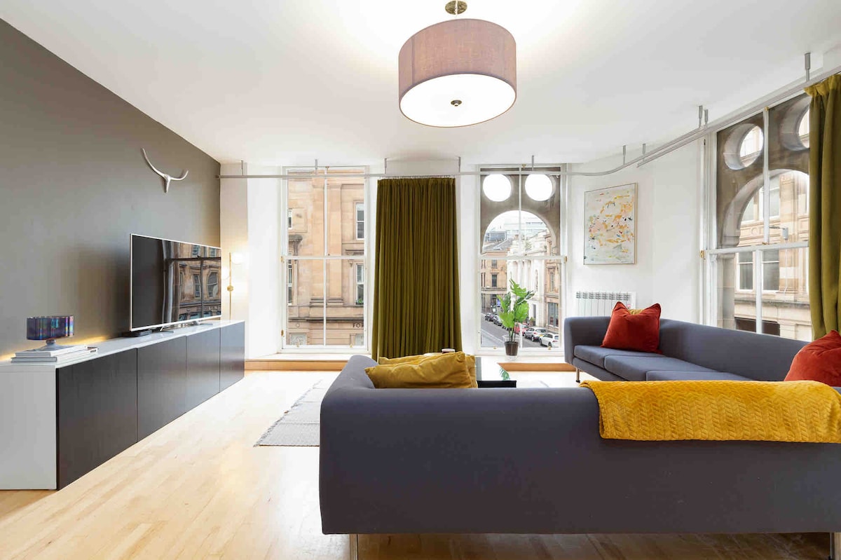 A modern living area is highlighted by large windows allowing natural light to fill the space. A dark sofa is complemented by colorful cushions, while a sleek TV is positioned on a contemporary console. Decorative wall art and greenery add vibrancy to the room.