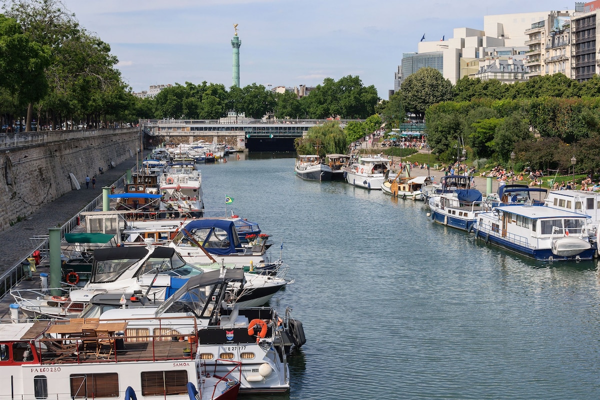 The image depicts a serene view of boats docked along a calm canal. Lush greenery lines the banks, with trees providing shade to pedestrians. In the background, a prominent monument stands visible, enhancing the urban landscape. The scene captures a tranquil waterway, evoking a leisurely atmosphere.