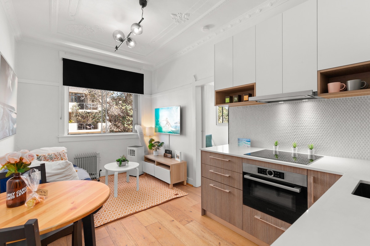 The kitchen and living area feature a modern design with a bright color palette. A well-equipped kitchen is visible, including sleek cabinetry and contemporary appliances. A small dining table sits beside a window, while a TV mounted on the wall faces a comfortable seating area, creating an inviting space.