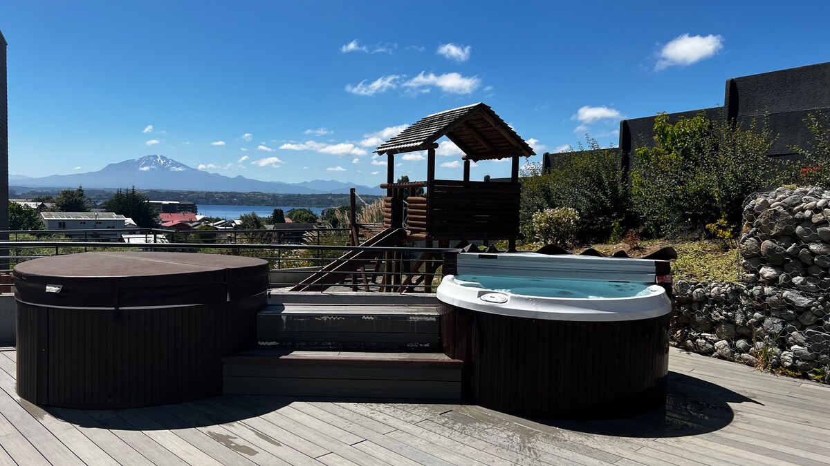 An outdoor area features two hot tubs, one circular and one rectangular, set against a backdrop of mountains and blue skies. Nearby, a wooden structure offers shade, enhancing the inviting atmosphere of relaxation. The deck is made of light-colored wood, complementing the natural scenery.