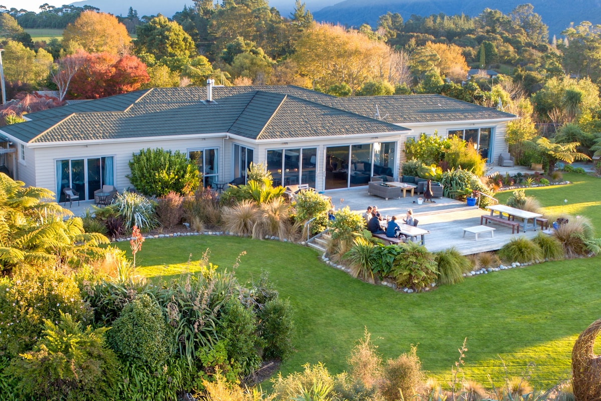 The exterior of Zatori is depicted, showcasing a spacious house surrounded by lush gardens and trees in autumn colors. A large deck extends from the house, featuring seating areas for relaxation. The panoramic view encompasses the well-maintained lawn and vibrant landscaping.