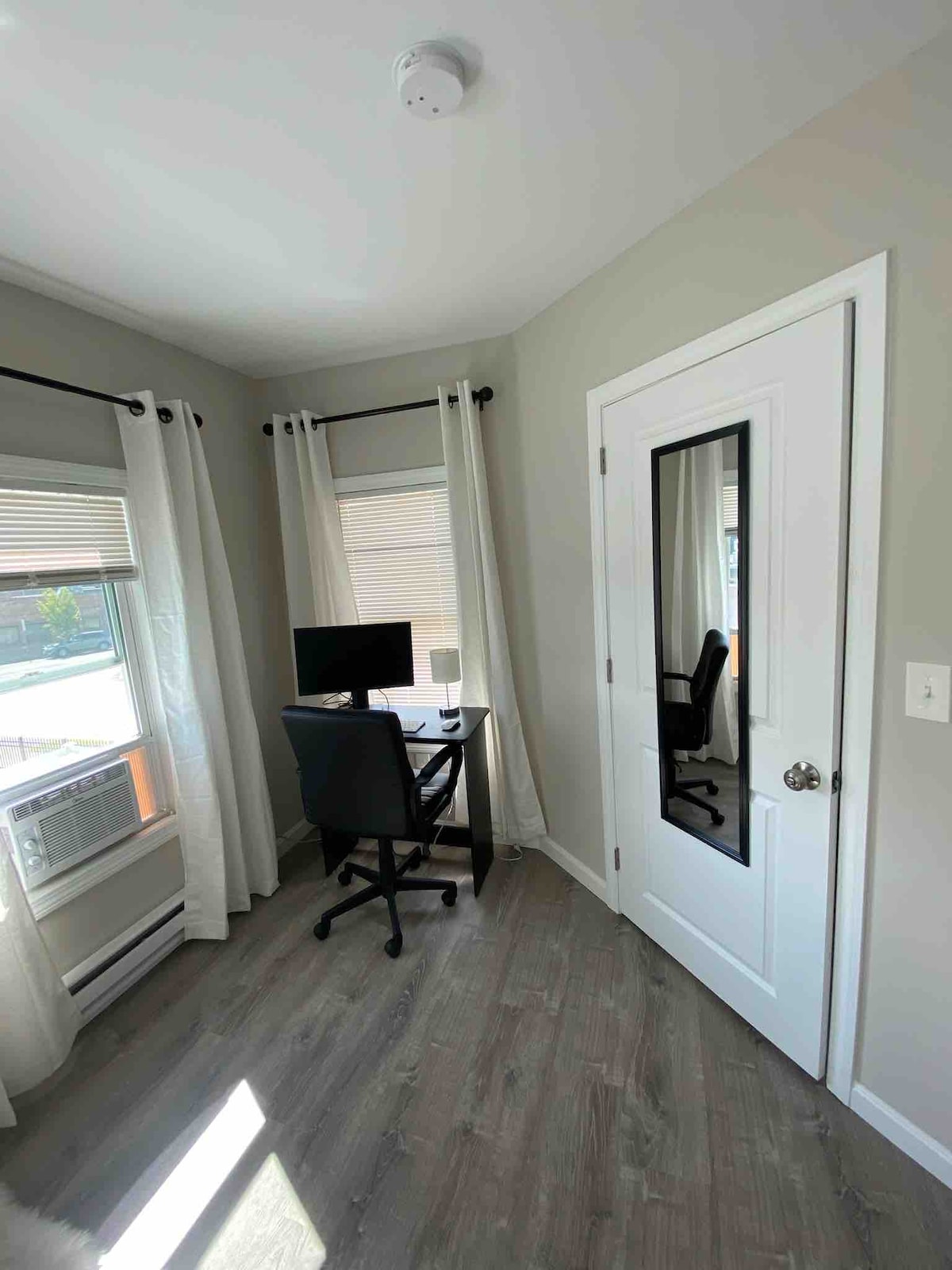 A dedicated workspace is featured in a well-lit corner of the apartment. A computer monitor, mouse, and keyboard are positioned on a small desk. Two comfortable chairs are present, with natural light streaming through the large window, framed by sheer curtains.