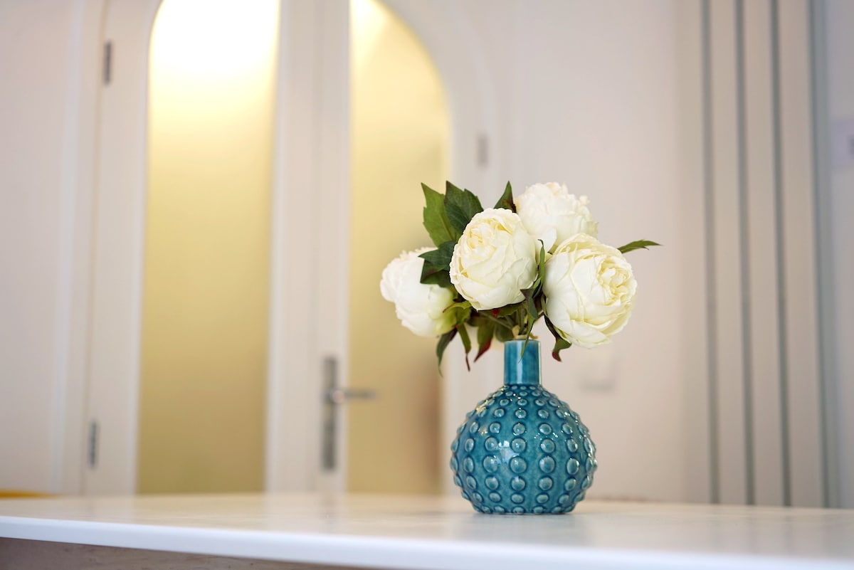 A simple, vibrant blue vase is placed on a white countertop, holding a bouquet of white peonies. In the background, an archway leads to a softly lit space, enhancing the freshness of the floral arrangement.