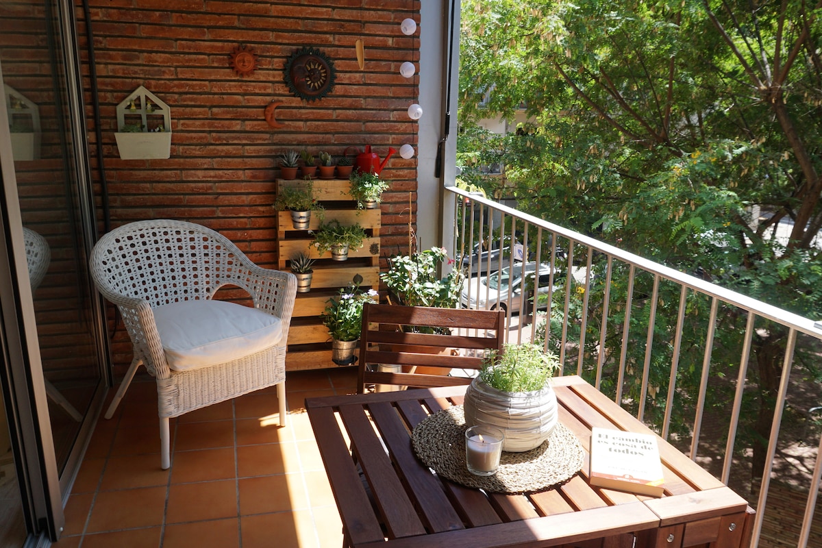 A charming balcony space features a small wooden table and a white wicker chair. Behind the table, a vertical garden displays various plants. The tiled floor and surrounding greenery create a serene environment, inviting relaxation while enjoying the view.