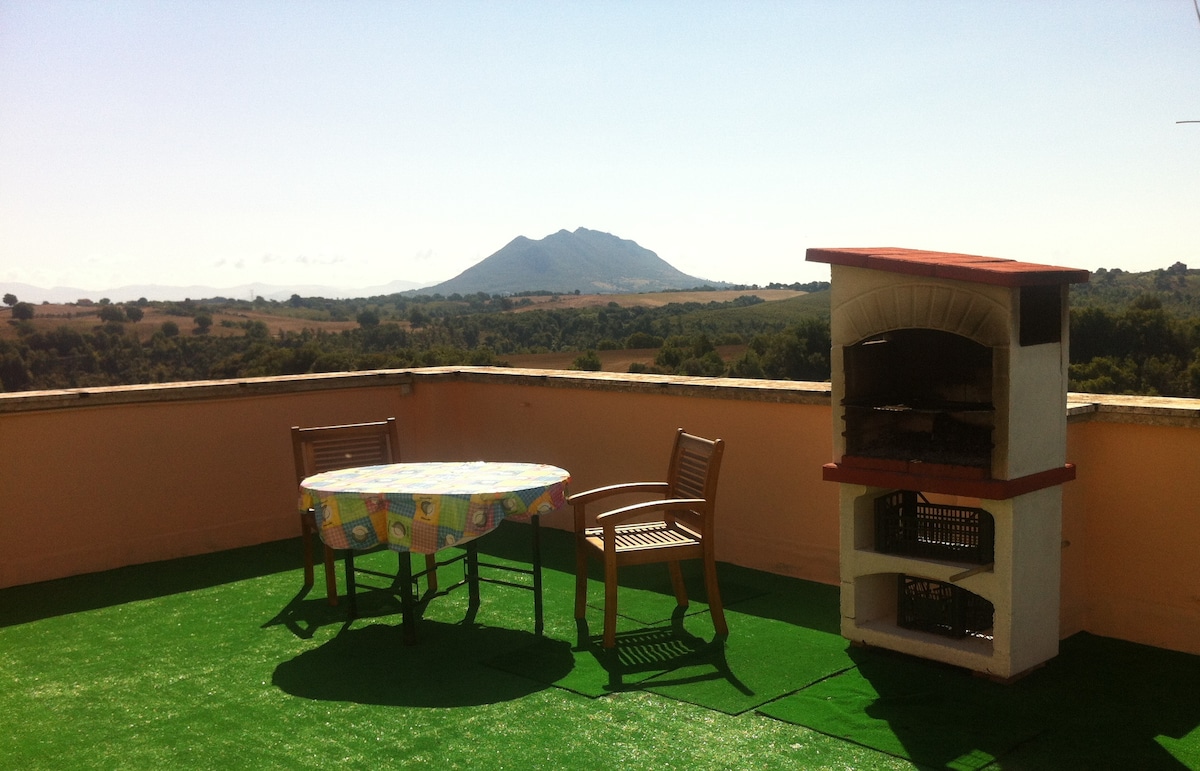 A large panoramic terrace features a round table with a colorful tablecloth and two wooden chairs, complemented by a stone barbecue. A scenic view of Mount Soratte is visible in the background, surrounded by green hills under a clear blue sky.