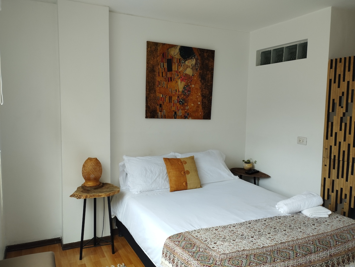 A comfortable bedroom features a spacious bed adorned with white bedding and an accent pillow. A piece of art hangs above the bed, and two nightstands flank it. Natural light enters through a nearby window, enhancing the simple and calming design.