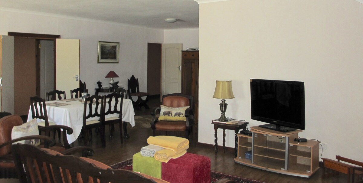 A spacious living area is presented with dark wooden furniture, including a comfortable seating arrangement and an entertainment unit featuring a television. A dining table with elegant chairs is visible, complemented by warm lighting from a nearby lamp. Fresh towels are neatly arranged on a decorative rug.