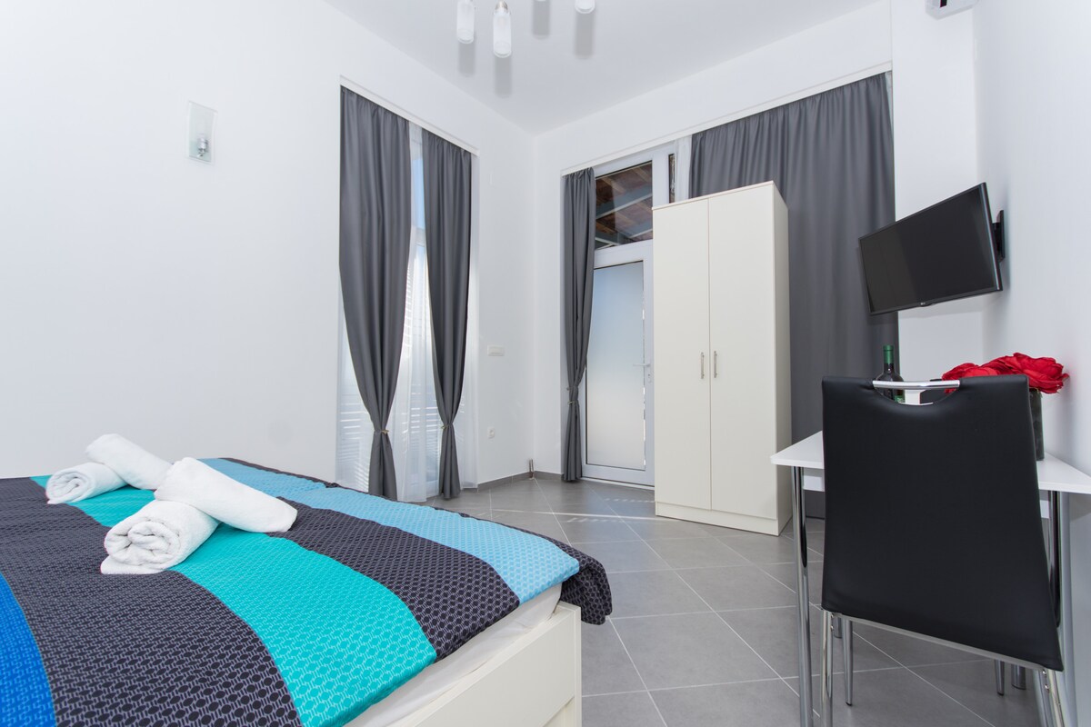 The bedroom features a spacious bed with colorful bedding and neatly arranged towels. A wardrobe stands adjacent, and a desk with a chair is positioned nearby. Natural light enters through a window, framed by gray curtains, adding to the room's bright and open feeling.
