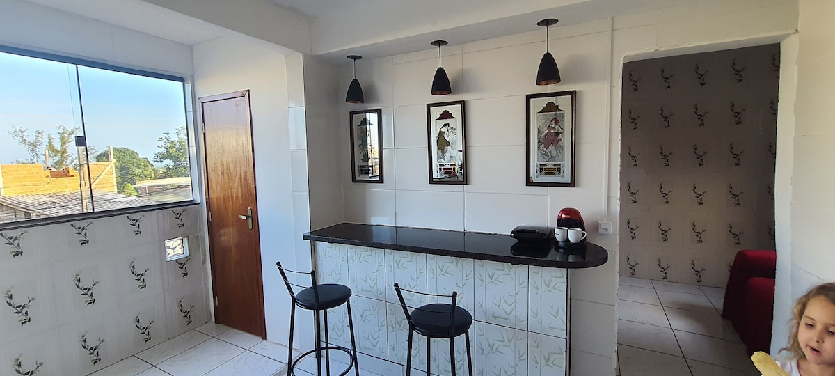 A bright kitchen area features a sleek black countertop with two high-backed stools positioned nearby. Wall-mounted artworks enhance the space, while pendant lights hang above. A large window allows natural light to fill the room, and a doorway leads to a different area of the apartment.