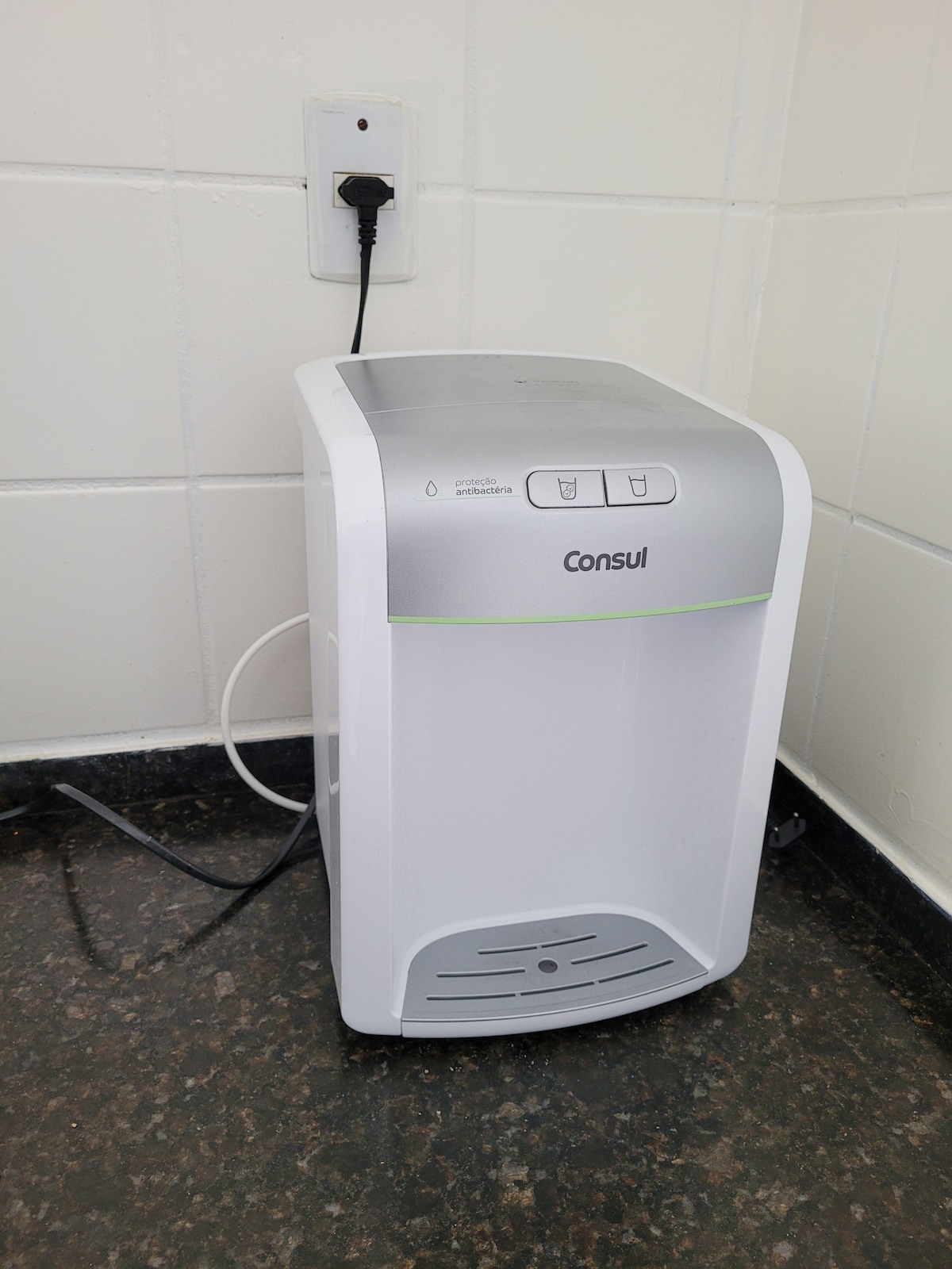 A dehumidifier is positioned against a wall, featuring a sleek white design with a silver top. Buttons for operation are visible on the front, and a power cord connects it to an electrical outlet above the countertop.