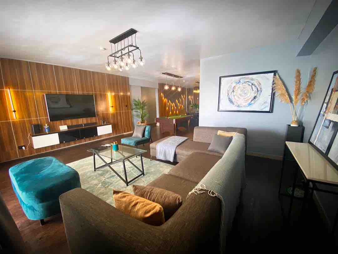A modern living area is highlighted by a stylish sectional sofa and a contrasting plush ottoman. A sleek entertainment unit is mounted against a wooden accent wall, while a vibrant piece of artwork adds a pop of color. Ambient lighting creates a cozy setting.
