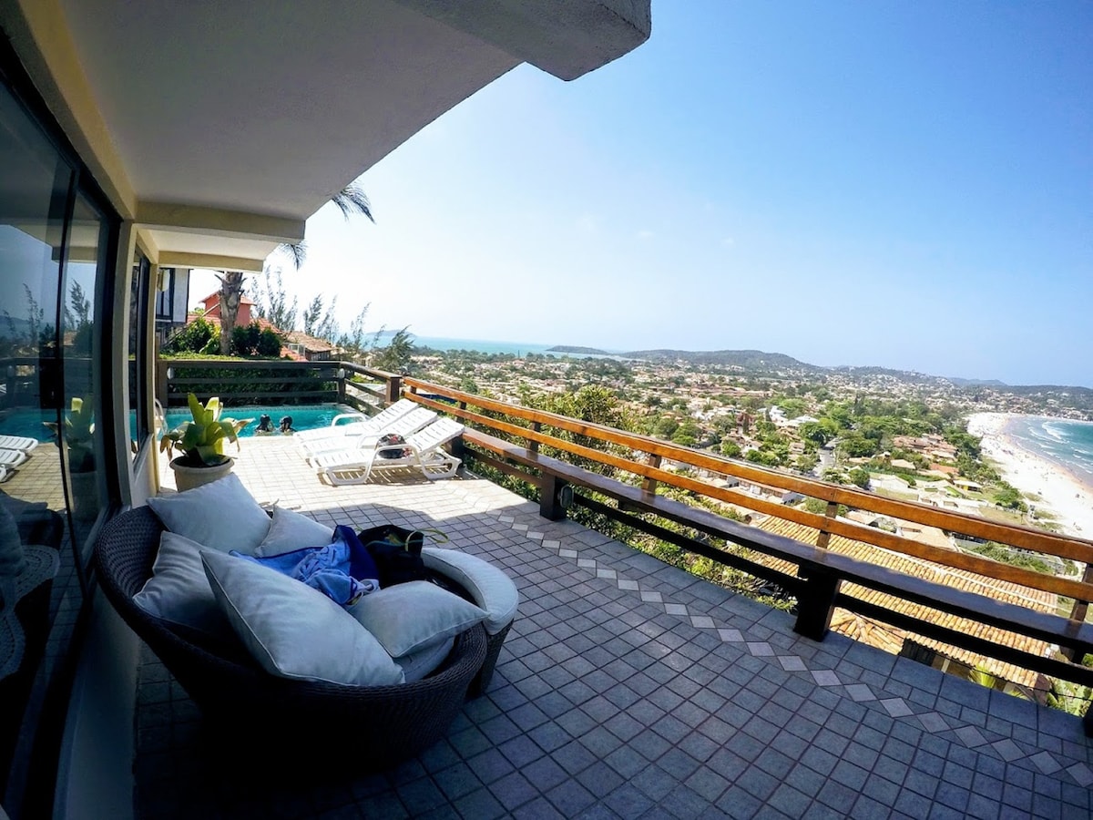A spacious outdoor terrace features a comfortable lounging area with plush cushions and a stunning view of the ocean and surrounding coastline. A private pool is visible nearby, framed by a wooden railing, creating an inviting space for relaxation.