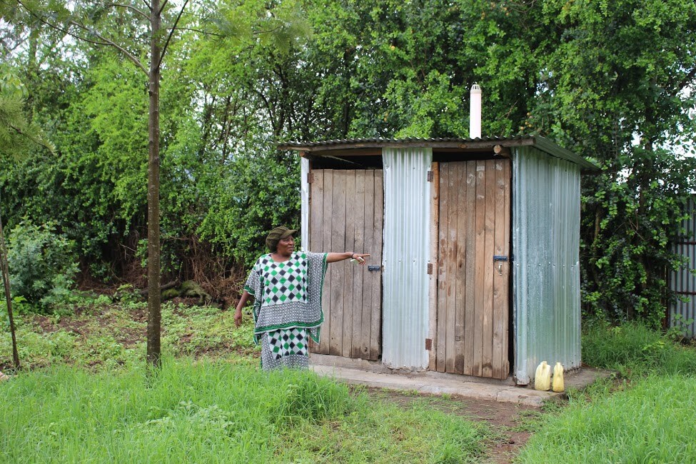 A simple outdoor shower and toilet block is shown, constructed with wooden and metal materials. The structure features two doors, while a person stands beside it, pointing towards the block. Surrounding greenery and a tree add to the natural setting.