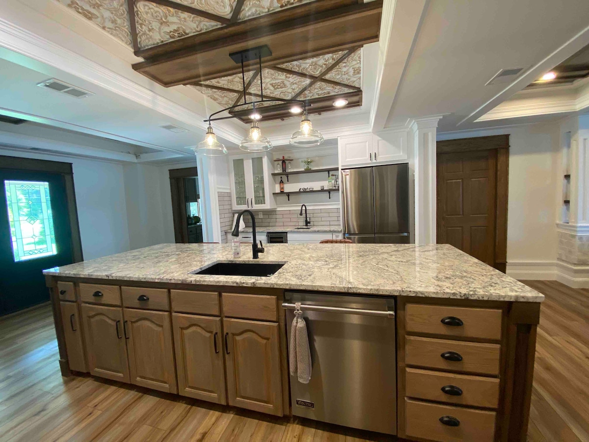 A spacious kitchen features a large granite island with a deep sink and polished wood cabinetry. Modern stainless steel appliances, including a refrigerator and dishwasher, are visible. Stylish pendant lights hang above the island, and a decorative ceiling adds architectural interest.