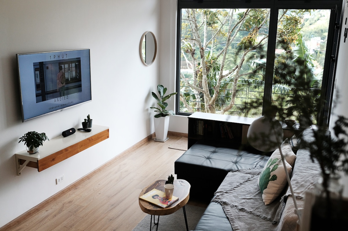 A living room offers a large window with views of the surrounding greenery. A 49-inch TV is mounted on one wall above a simple wooden shelf. A comfortable, dark sofa and round coffee table, adorned with a drink and a plant, create a welcoming environment.