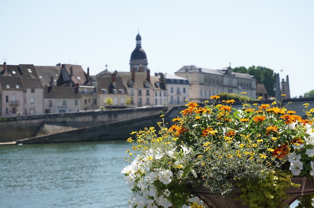 A vibrant flower arrangement in the foreground features a mix of yellow, orange, and white blooms. In the background, a serene river flows alongside historical buildings, with a prominent dome and bell tower visible against a clear blue sky.