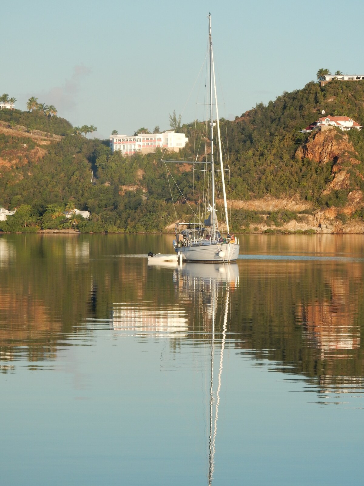 A sleek 50-foot sailboat is anchored in calm waters, reflecting its image on the surface. Surrounding hills display verdant greenery, with a hint of residential buildings perched above. The serene landscape suggests tranquility and connection with nature.