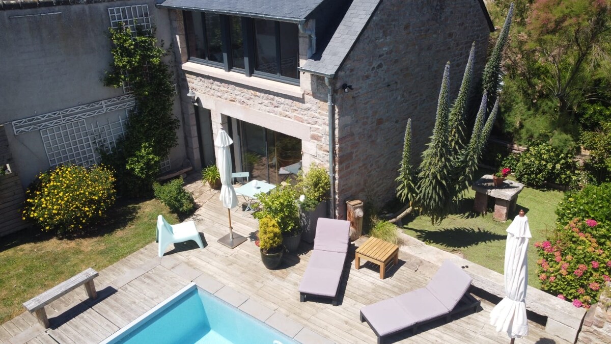 An aerial view showcases a charming stone house with large glass doors leading to a pool area. Sun loungers and umbrellas surround the heated pool. Lush greenery and colorful flowers adorn the garden, enhancing the outdoor space for relaxation.
