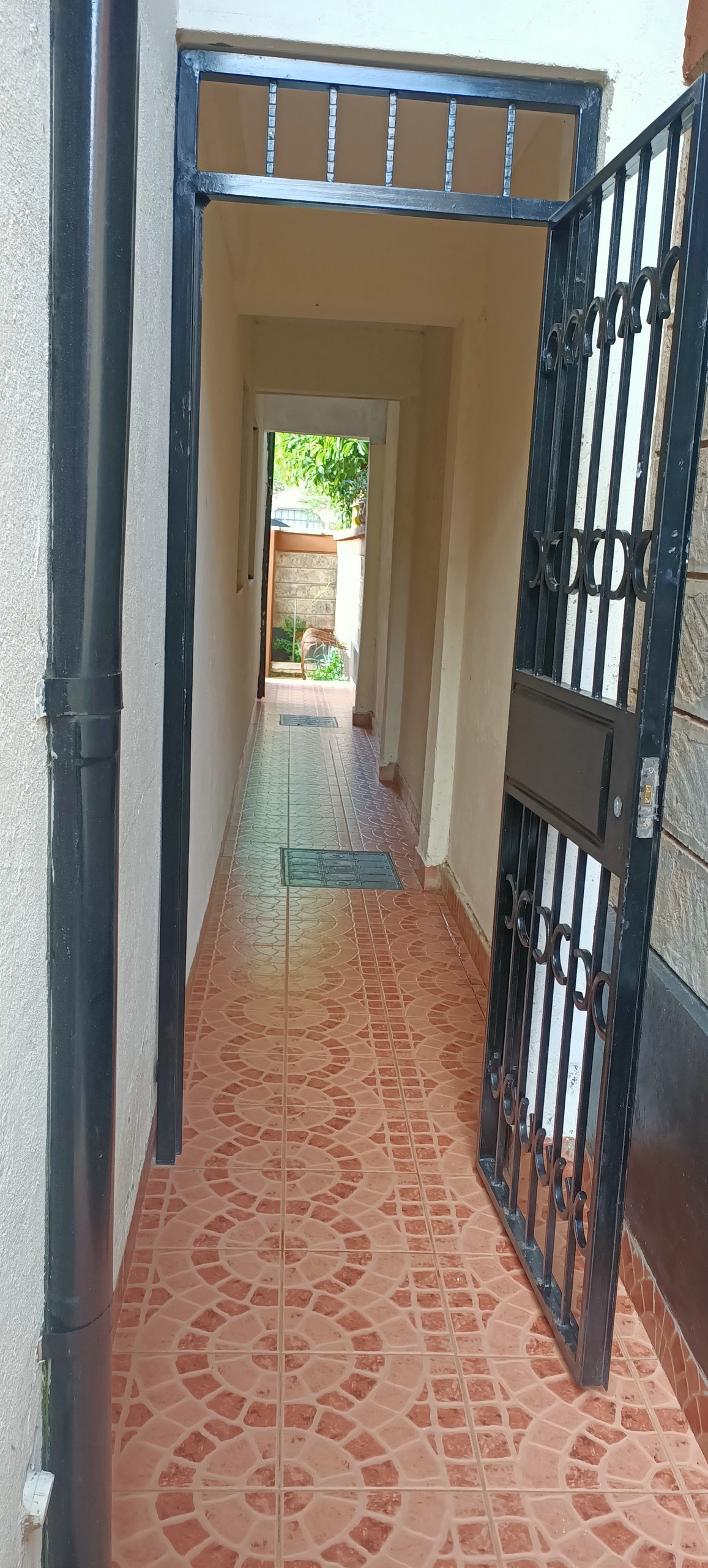A narrow hallway is visible, featuring a tiled floor and bordered by light-colored walls. A gate stands at the entrance, and natural light filters in from nearby openings, illuminating the space. A mat is placed at the foot of the entrance.