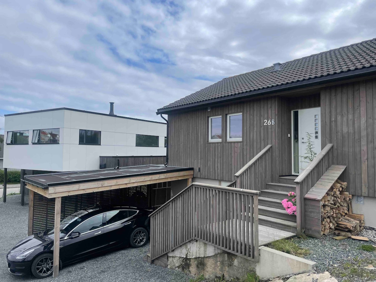 A wooden structure features a covered carport for vehicle parking, with a sleek black car parked below. Wooden steps lead to the entrance, framed by a small flower arrangement. The exterior showcases a mix of natural materials and a modern nearby building.