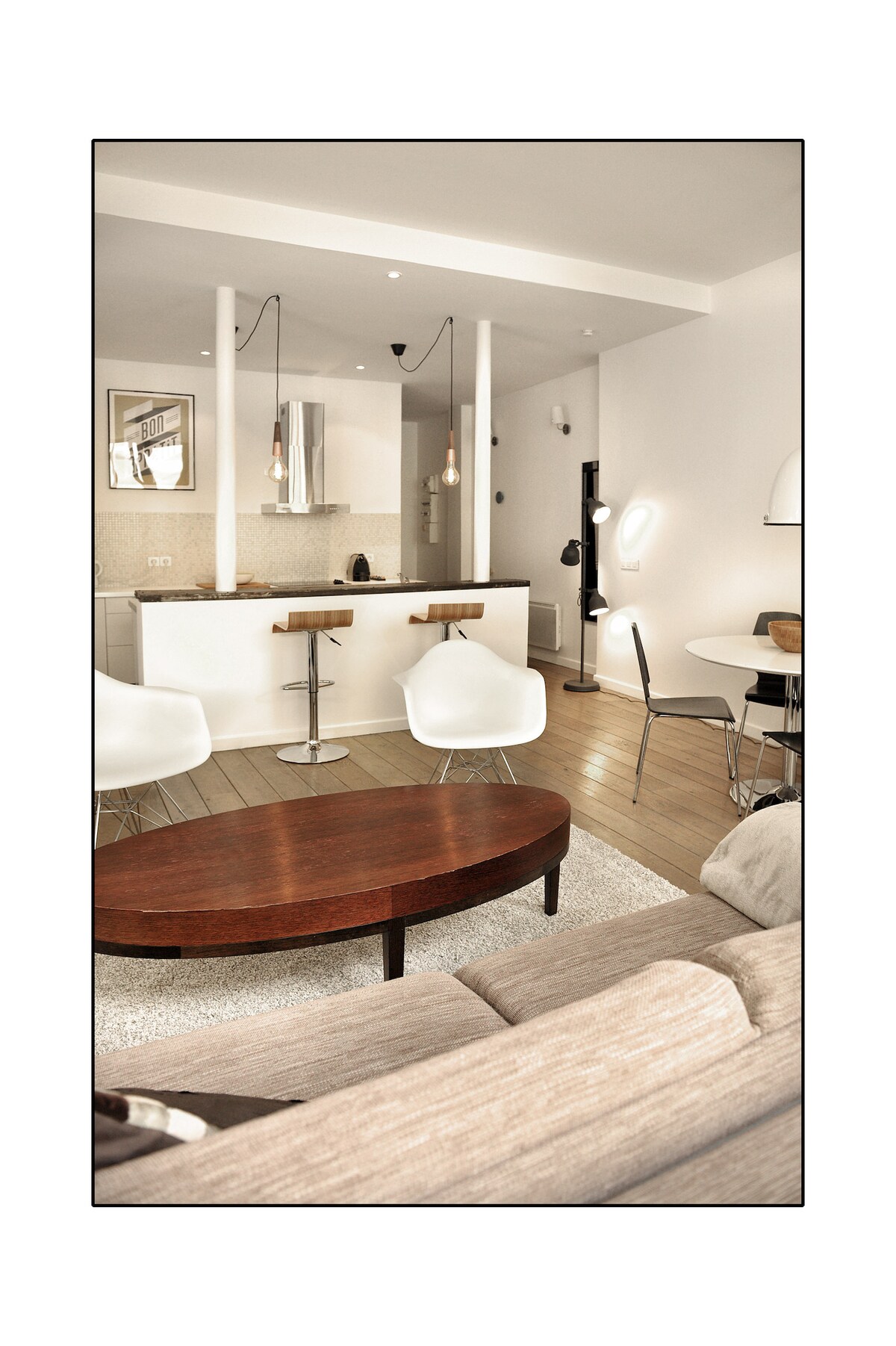 A modern living space features a comfortable sofa with soft cushions, accompanied by two white chairs and a circular wooden coffee table. A dining area with a round table is visible, and the open kitchen displays sleek appliances, enhancing the airy atmosphere of the room.