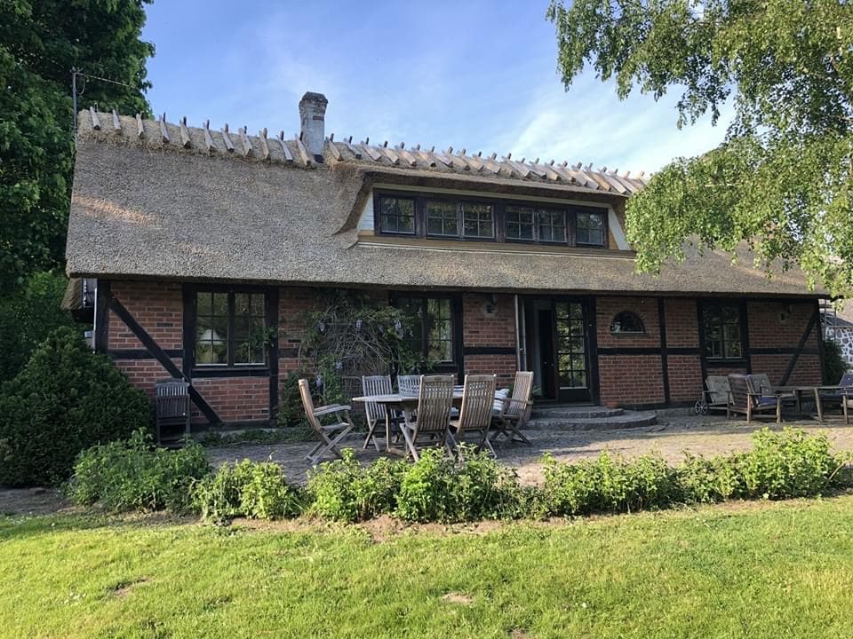 A charming house with a thatched roof is set within a lush garden. Outdoor seating is arranged around a large table on a stone patio, surrounded by greenery. Tall trees provide shade, and the sky is clear, enhancing the inviting atmosphere of the outdoor space.