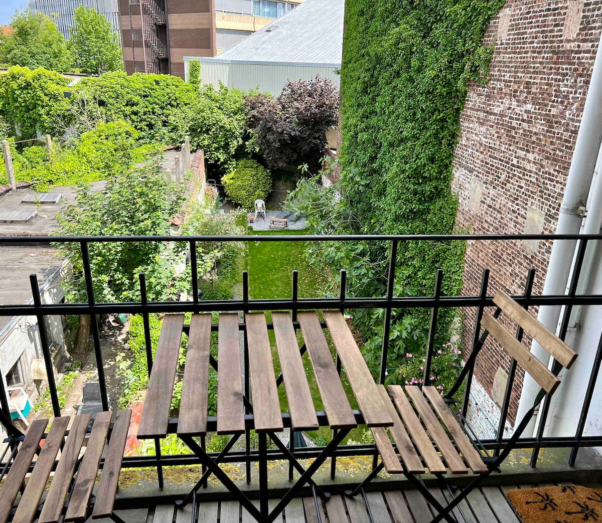 A small balcony overlooks a lush garden area filled with greenery and trees. Two wooden folding chairs are positioned at the edge of the balcony, providing a serene view of the outdoor space below.