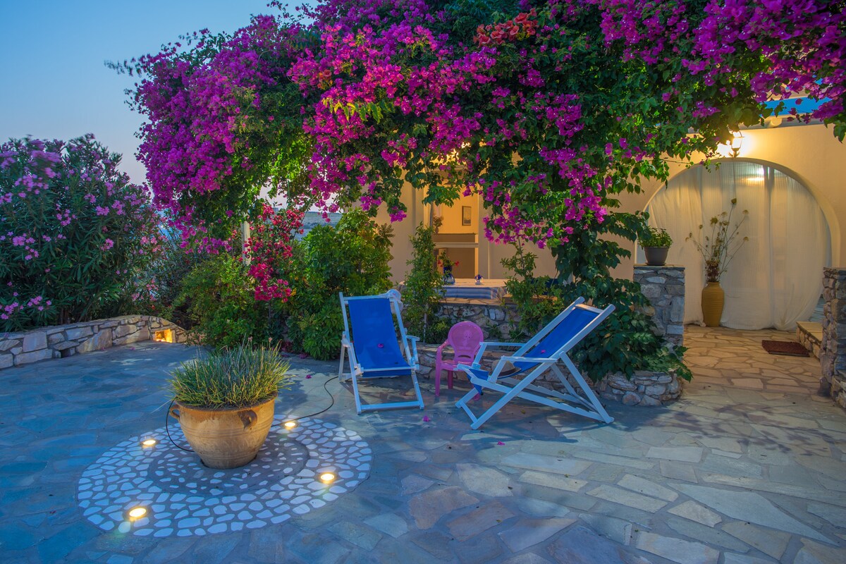 A charming outdoor area is surrounded by vibrant bougainvillea and other greenery. Two lounge chairs are positioned on a stone patio, complemented by a decorative planter at the center. Soft lighting creates a welcoming atmosphere, perfect for relaxation or enjoying the surroundings.