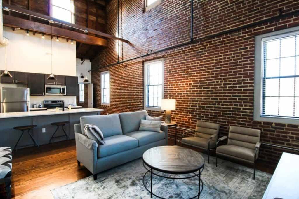 A spacious living area features exposed brick walls and hardwood flooring. A comfortable gray sofa is situated across from a round coffee table. Two contemporary chairs complement the seating arrangement, while natural light enters through multiple windows, illuminating the room.